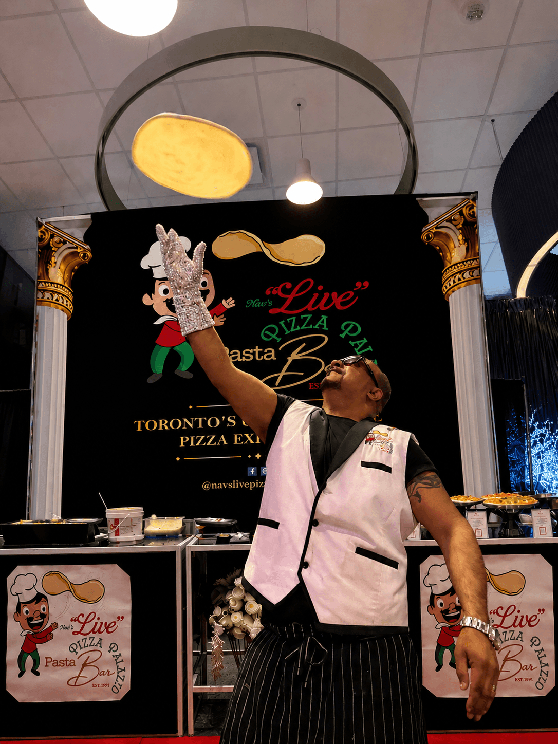 Nav tossing pizza dough in the air wearing a crystal glove in front of the Live Pizza Palazzo trade-show backdrop