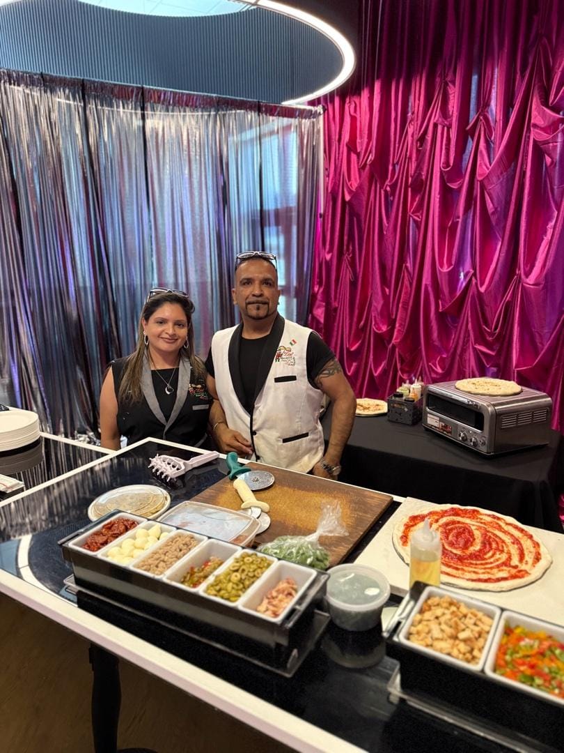 Nav and a team member behind the live prep station with fresh toppings and saucing a pizza base