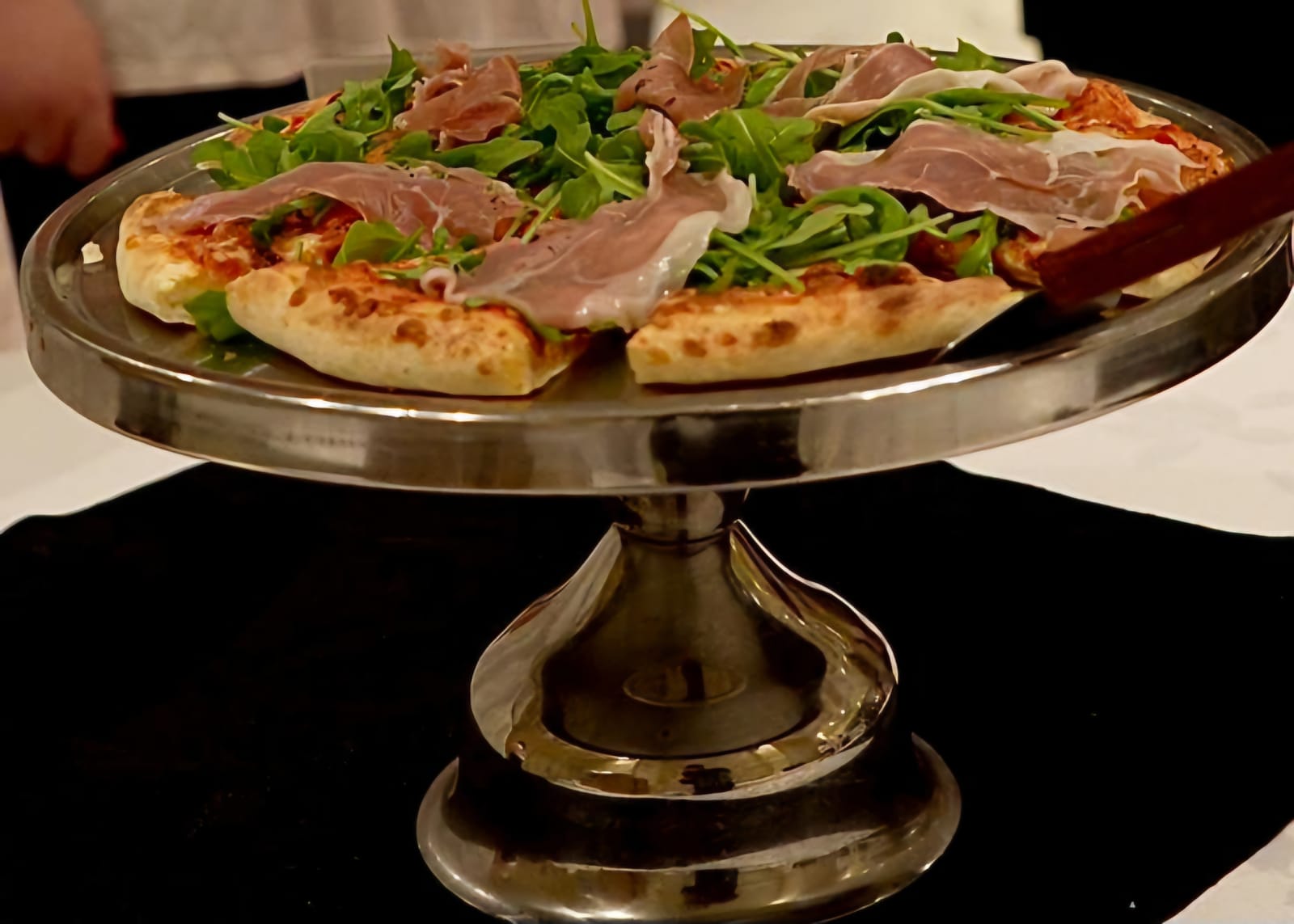 Prosciutto and arugula pizza topped with cured ham and fresh greens on a silver stand