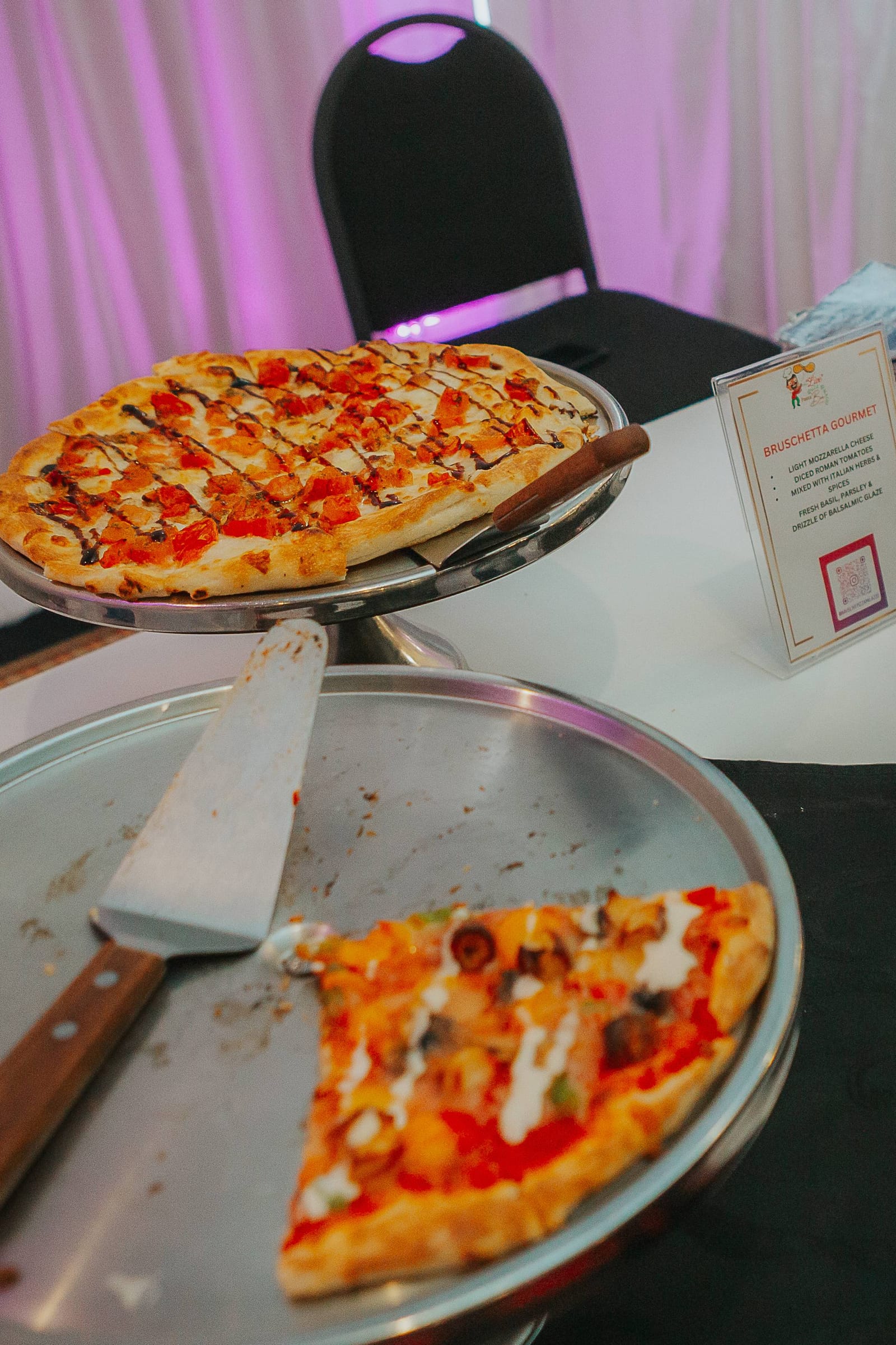 Bruschetta gourmet pizza on a silver stand with a menu card under purple draping at a wedding